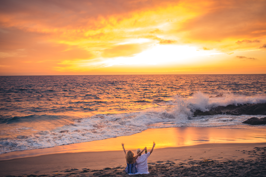 couple sitting at the beach, watching the sunset, holding hands up, happily 